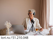 Elderly eastern woman hosting podcast in cozy home with laptop and microphone. Стоковое AI-изображение, AI-художник Евгения Пархоменко / Фотобанк Лори