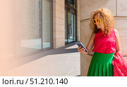 Coquettish s young female model in fashionable elegant bright multicolored green and red clothes looks at her phone against the background of the columns of an office building in the city. Стоковое фото, фотограф Anastasia Kayander / Фотобанк Лори