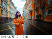 Молодая женщина с фиолетовыми волосами в оранжевом дождевике стоит под прозрачным зонтом на мокрой городской улице. Стоковое AI-изображение, AI-художник Кирилл Сиротюк / Фотобанк Лори