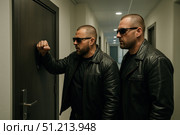 Two Vigilant Security Agents in Black Leather Jackets Knock on a Door, Ready for Action in a Modern, Dimly Lit Hallway Environment. Стоковое AI-изображение, AI-художник Башкирова Анна / Фотобанк Лори