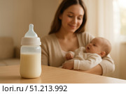 A Cozy Moment: A Mother Holding Her Baby with a Bottle of Milk on the Table, Embracing the Warmth of Family and Nurturing Love in Their Home Environment. Стоковое AI-изображение, AI-художник Башкирова Анна / Фотобанк Лори