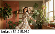 Happy woman dancing in cozy home interior with many plants, beautiful carefree female in white floral dress enjoying slow living for lifestyle or wellbeing concept. Стоковое AI-изображение, AI-художник Павел Муравьев / Фотобанк Лори