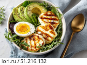 Grilled Halloumi Cheese and avocado with a runny egg in a bowl black pepper sprinkled on top. Halloumi breakfast served with a golden spoon, on a light blue linen cloth. Delicious-looking dish. Стоковое AI-изображение, AI-художник Марина Демидюк / Фотобанк Лори