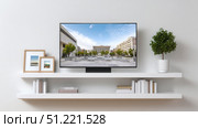 Large modern television displaying vibrant cityscape, positioned on white wall shelf among books, frames, potted plant, presenting contemporary living room design. Стоковое AI-изображение, AI-художник Юрий Поздников / Фотобанк Лори