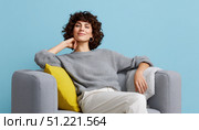 Curly haired woman wearing gray sweater and white pants lounging comfortably on gray sofa, soft yellow pillow and blanket adding warmth to peaceful home setting. Стоковое AI-изображение, AI-художник Юрий Поздников / Фотобанк Лори