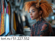 A young woman with curly red hair sits at a computer, working intently on a project. She is wearing a stylish sweater with an abstract pattern in orange and black tones, and her unique earrings add personality to the image. A bright fragment of fabric or a designer pattern is visible on the monitor screen, indicating work in the field of fashion, graphic design or creativity. The background is slightly blurred, creating an emphasis on the model and her focused gaze. The atmosphere of the photo conveys inspiration, creativity and concentration on work. Стоковое AI-изображение, AI-художник Анна Кривицкая / Фотобанк Лори