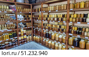 A honey shop where jars with different varieties of this natural product are neatly displayed on wooden shelves. Glass jars of different sizes — from small 0.25 l to large three-liter ones — are filled with honey of all possible shades: from almost white to dark brown. The varieties are visible: buckwheat, linden, sweet clover, hawthorn and others. There are price tags under each row. The interior is dominated by wooden shelves, creating an atmosphere of home comfort. On some shelves you can also see nuts in honey, herbal infusions and souvenirs, which gives the room the flavor of a farmer's shop. Стоковое фото, фотограф Анна Кривицкая / Фотобанк Лори