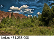 A landscape with a bright green summer forest and thick grass in the foreground. A brownish hill or mountain rises on the horizon, creating a contrast with the green of the trees. A rich dark blue sky stretches above the landscape, decorated with voluminous white clouds, creating a dramatic and picturesque effect. The combination of bright greenery, brown rocks and deep sky tones makes the picture expressive and dynamic. This shot is suitable for nature, ecology, travel and landscape photography. Стоковое фото, фотограф Анна Кривицкая / Фотобанк Лори