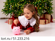 Banking, finances, investment and happy people concept. Cute happy little girl lies a floor and puts money coins in pink piggy bank on Christmas. Pocket money, family budget and savings concept. Стоковое фото, фотограф Ирина Михайличенко / Фотобанк Лори