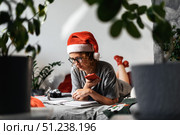 Woman in Santa hat relaxes at home during Christmas holiday, using smartphone to enjoy winter moments. Surrounded by cozy decor, she connects through technology, celebrating Christmas digitally. Стоковое фото, фотограф Енваев Андрей / Фотобанк Лори