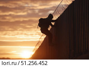 A silhouette of an individual climbing a barbed-wire fence during sunset, symbolizing the challenges of migration and borders in a dramatic landscape. Стоковое AI-изображение, AI-художник Башкирова Анна / Фотобанк Лори