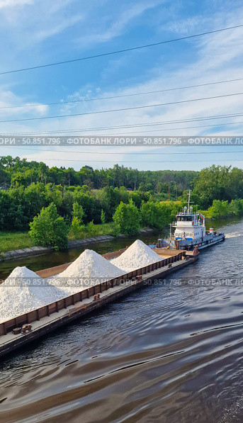 A barge with a cargo of white rubble is moving along the Volga River, which is being dragged by a small tugboat with a Russian flag. Green trees and sparse woodlands are visible on the shore. Стоковое фото, фотограф Анна Чижова / Фотобанк Лори
