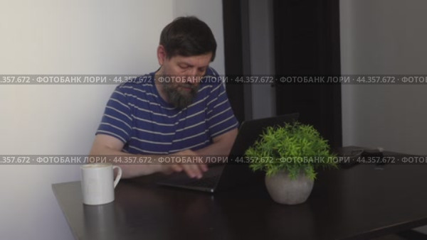 A bearded man is working on a laptop in the kitchen at a black table. Стоковое видео, видеограф Ярославский Максим / Фотобанк Лори