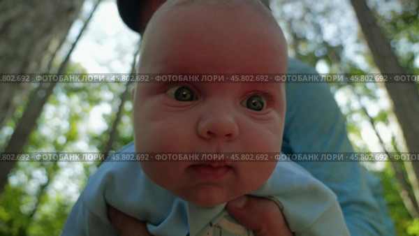 A bearded man tilts a baby in a blue onesie towards the camera until the baby's face takes up the entire frame. Sunlit forest in the background. Father and son in nature. Стоковое видео, видеограф Воловодов Владимир / Фотобанк Лори