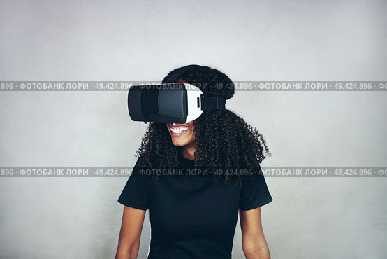 A beautiful young black woman with curly afro hair wears virtual reality VR headset and plays videogames while smiling in studio with grey background. Стоковое фото, агентство Ingram Publishing / Фотобанк Лори