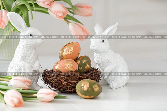 A bouquet of delicate tulips, Easter bunnies and eggs with a golden pattern on the table. There is a white kitchen in the background. Beautiful greeting card. The concept of minimalism. Стоковое фото, фотограф Photojuli / Фотобанк Лори