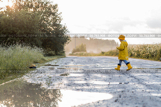 A boy in yellow rubber boots and a raincoat runs through puddles, having fun and playing with a remote-controlled car after it rains. Summer and autumn holidays. A happy childhood. Стоковое фото, фотограф Photojuli / Фотобанк Лори