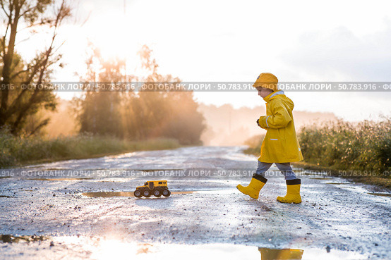 A boy in yellow rubber boots and a raincoat runs through puddles, having fun and playing with a remote-controlled car after it rains. Summer and autumn holidays. A happy childhood. Стоковое фото, фотограф Photojuli / Фотобанк Лори