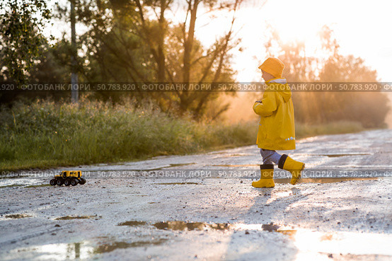 A boy in yellow rubber boots and a raincoat runs through puddles, having fun and playing with a remote-controlled car after it rains. Summer and autumn holidays. A happy childhood. Стоковое фото, фотограф Photojuli / Фотобанк Лори
