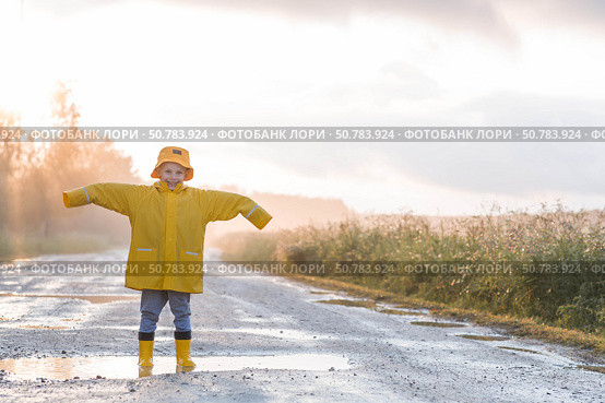 A boy in yellow rubber boots and a raincoat runs through puddles, having fun and playing after it rains. Summer and autumn holidays. A happy childhood. Стоковое фото, фотограф Photojuli / Фотобанк Лори