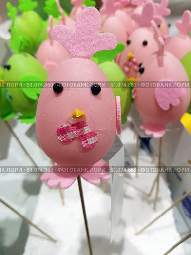 A bright decorative figurine in the form of a chicken, made in pink tones. The base is a soft pink plastic egg, decorated with eyes made of black beads, a small yellow beak and a pink fabric checkered bow. A comb made of pink felt is attached to the top, and wings and legs made of the same material are at the bottom and on the side. The figurine is mounted on a wooden skewer, which allows it to be used as a festive decoration or an element of the composition. In the background, other similar figurines in pink and green colors are visible, slightly blurred, which creates the effect of depth. Стоковое фото, фотограф Анна Кривицкая / Фотобанк Лори