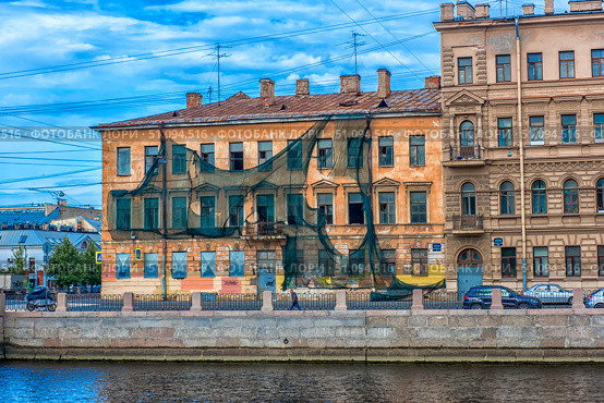 A building in the historic center of Saint Petersburg, currently undergoing renovation or showing signs of neglect. The facade is draped with protective netting, some windows are broken, and the plaster is deteriorating. In the foreground is an embankment and a canal or river. The image captures the contrast between cultural heritage and the passage of time — a typical scene in the older districts of Saint Petersburg, rich with charm and historic character. (2017 год). Редакционное фото, фотограф Анна Кривицкая / Фотобанк Лори