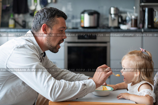 A caring man with a beard feeds his little daughter. A little girl sits at a children's dining table and dines with her dad. Happy fatherhood. Стоковое фото, фотограф Михаил Решетников / Фотобанк Лори