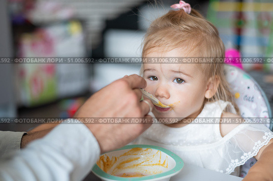 A caring man with a beard feeds his little daughter. A little girl sits at a children's dining table and dines with her dad. Happy fatherhood. Стоковое фото, фотограф Михаил Решетников / Фотобанк Лори