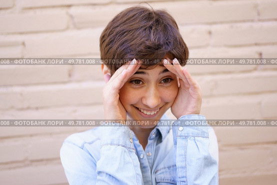 A cheerful individual with short hair shows surprise and joy, playfully placing their hands on their head against a warm beige brick background that enhances the mood of happiness. A Joyful Young Person Expressing Genuine Surprise and Delight in a Vibrant Setting. Стоковое фото, агентство Ingram Publishing / Фотобанк Лори