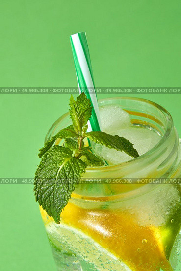 A close-up of a green mint in focus in a glass jar with citrus slices , ice, sparkling water and colored plastic straw isolated on a green background. Natural cold summer homemade lemonade.. A sprig of green mint in focus in a glass jar with a cold natural homemade lemonade isolated on green. Citrus fruits slices of lemon and lime, ice, water and plastic straw in the glass. Стоковое фото, агентство Ingram Publishing / Фотобанк Лори