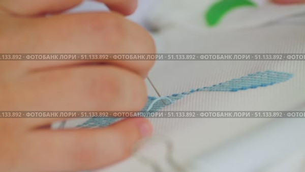 A close-up of the process of cross-stitch embroidery. A hand delicately guides the needle with blue thread through the white fabric. The complex art of embroidery. Стоковое видео, видеограф Воловодов Владимир / Фотобанк Лори