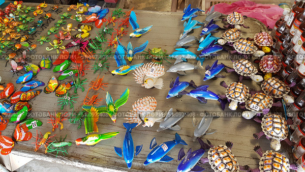 A counter with bright souvenirs in the form of animals and insects. On the wooden surface are laid out multi-colored figures of butterflies, fish, parrots, dolphins, turtles, crabs, octopuses and other sea and land creatures. The toys are made of plastic or metal and painted in rich colors - red, green, blue, yellow, orange. Some of them have price tags with numbers. In the right part of the frame, some bottles with liquid are visible, probably from the neighboring counter. The atmosphere is reminiscent of an oriental market or a tourist souvenir shop. Стоковое фото, фотограф Анна Кривицкая / Фотобанк Лори