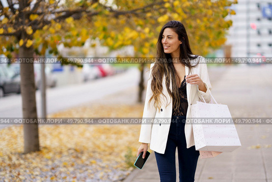 A fashionable woman enjoys a delightful autumn shopping day, confidently holding colorful bags in one hand and a smartphone in the other, savoring every joyful moment of her outing. A fashionable woman joyfully shops among colorful autumn leaves using her smartphone. Стоковое фото, агентство Ingram Publishing / Фотобанк Лори