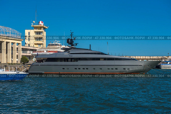 A fragment of the seaport in Yalta. In the center of the frame, an elegant and modern yacht called Orion is moored, painted dark gray with an orange line along the waterline. Nearby are boats and other vessels, and in the background are visible buildings of the port infrastructure, including the control tower with a characteristic dome. The sea is bright and rich blue, light ripples on the water indicate a fresh breeze. On the concrete wall behind is the inscription WELCOME, which emphasizes the tourist character of the place.The photo conveys the atmosphere of active marine life and the modern Yalta coast. (2021 год). Редакционное фото, фотограф Анна Кривицкая / Фотобанк Лори