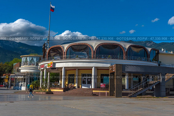 A fragment of the Yalta embankment, one of the most popular resort towns in Crimea. In the foreground is a modern building with a round façade and stained-glass windows, where cafes and restaurants are located. On the left is part of the pedestrian zone with passers-by, vacationers and strollers along the shore. In the background, the green mountains of the Crimean ridge rise, and the sky is decorated with living clouds and a flying bird. The Russian flag on the mast emphasizes the formality and maritime character of the place.The decor conveys an atmosphere of relaxation, resort activity and harmony with the natural landscape. (2021 год). Редакционное фото, фотограф Анна Кривицкая / Фотобанк Лори