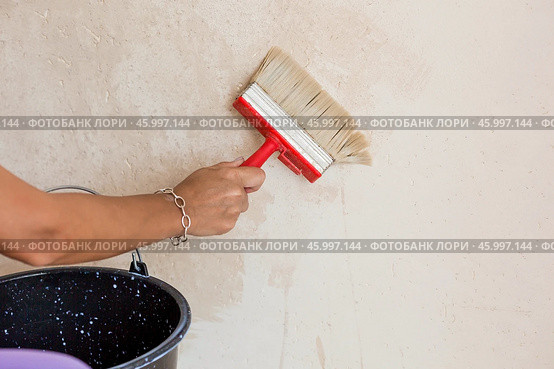 A hand with a wide brush and a bucket next to the wall being repaired during the application of primer or glue before gluing wallpaper. Wall treatment with a primer. Стоковое фото, фотограф Photojuli / Фотобанк Лори