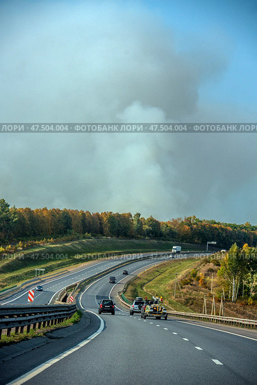 A highway stretching into the distance with a stream of cars moving along it. Behind the road, a dense forest is visible, from behind which a huge column of white-gray smoke rises, indicating a possible forest fire or a large blaze. The contrast between the clear road, greenery and the alarming cloud of smoke creates a sense of impending danger. The sky is partially covered with smoke. Стоковое фото, фотограф Анна Кривицкая / Фотобанк Лори