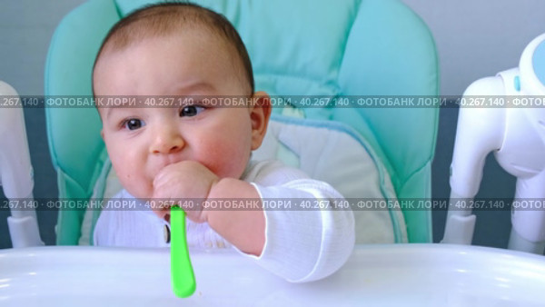 A hungry baby is gnawing on a plastic spoon at the table on a high chair. Teething, whims, itchy gums, introduction of complementary foods. Стоковое видео, видеограф Ольга Симонова / Фотобанк Лори