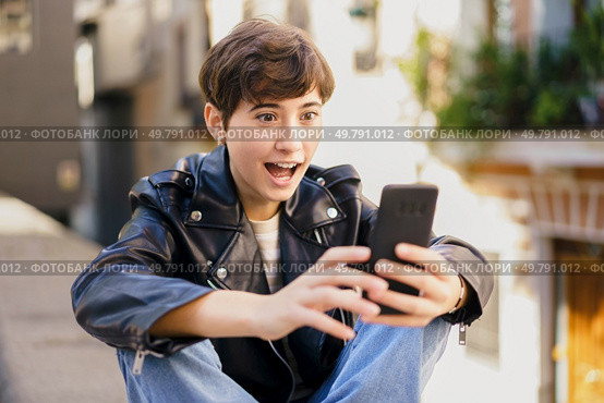 A joyful young person enthusiastically interacts with their smartphone while outdoors, vividly showcasing their excitement and connection in a lively and vibrant urban environment filled with energy. A cheerful young person is happily engaging with their smartphone in a bustling urban environment. Стоковое фото, агентство Ingram Publishing / Фотобанк Лори