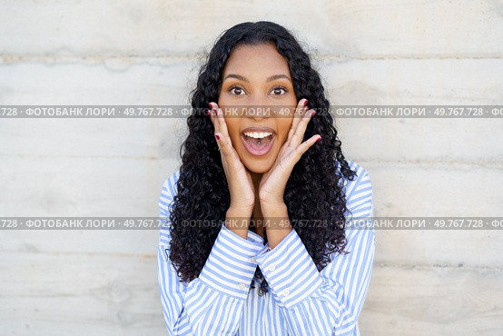 A joyful young woman with curly hair exclaims with excitement, showcasing her happiness against a neutral backdrop that highlights her vibrant personality and fills the air with warmth. An Excited Young Woman with Beautiful Curly Hair Expressing Joy and Enthusiasm Clearly. Стоковое фото, агентство Ingram Publishing / Фотобанк Лори
