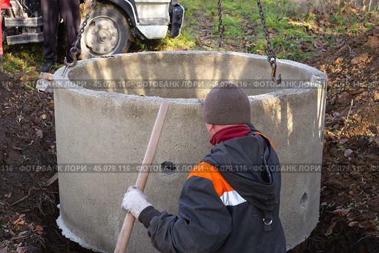 A loader lowers a concrete ring into a dug hole to build a septic tank. A worker installs a sewer into the ground. Стоковое фото, фотограф Коханова Наталья / Фотобанк Лори