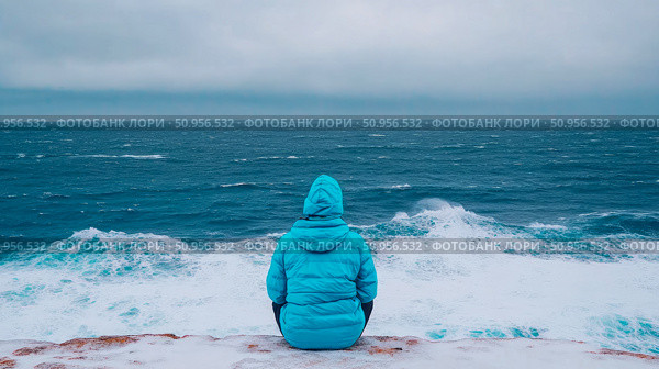 A lone figure in a vibrant blue jacket sits facing the vast, turbulent ocean under an overcast sky, contemplating the immense power of nature from a snowy cliff edge. Стоковое AI-изображение, AI-художник Данила Большаков / Фотобанк Лори