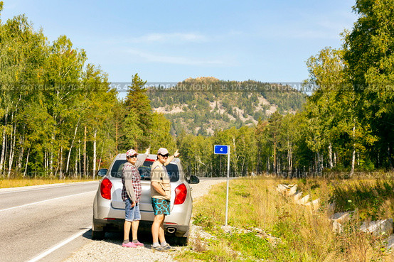 a man and a woman autotourists stand on the highway near the car and point to the high mountain Aleksandrovskaya Sopka on a sunny summer day. Стоковое фото, фотограф Акиньшин Владимир / Фотобанк Лори