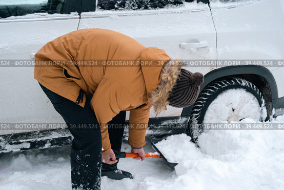 A man digs out a stalled car in the snow with a car shovel. Transport in winter got stuck in a snowdrift after a snowfall, sat on the bottom. First aid, tow truck, winter tires spikes and all-season. Стоковое фото, фотограф Ольга Симонова / Фотобанк Лори