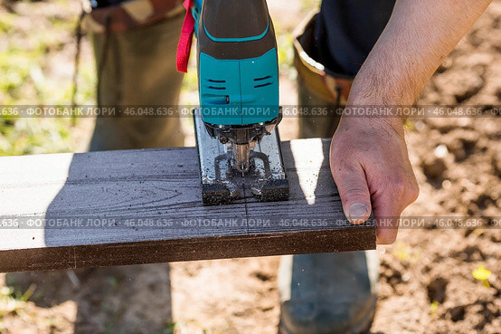 A man holds a battery-powered electric saw and builds new beds in a greenhouse made of wood-polymer composite. Macro photography. Side view. Preparing for the spring garden season. Стоковое фото, фотограф Photojuli / Фотобанк Лори