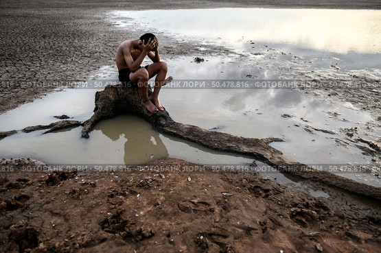 A man sat bent his knees and put his hands on his head, on the base of the tree and surrounded by water. Стоковое фото, агентство Ingram Publishing / Фотобанк Лори
