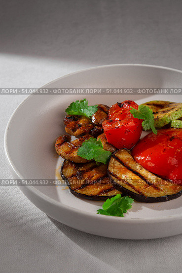 A mix of grilled vegetables including eggplant, zucchini and red bell pepper, garnished with fresh cilantro and served on a clean white ceramic plate. Стоковое фото, фотограф Aleksandr Gavrilychev / Фотобанк Лори