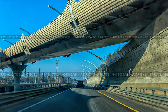 A modern section of a highway in St. Petersburg. The photo was taken from a car and shows an overpass with noise protection screens that rise above the road, creating a dynamic architectural perspective. In the foreground is a black car moving along perfectly smooth asphalt with yellow markings. On the sides are visible concrete fences, metal safety barriers and railway lines with power transmission line masts. In the background rises a red and white radio tower. The bright blue sky emphasizes modernity and open space. The atmosphere of the photo conveys the energy of the metropolis and the modern transport infrastructure of St. Petersburg. Стоковое фото, фотограф Анна Кривицкая / Фотобанк Лори