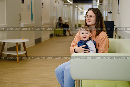 A mother woman with a toddler baby on a couch in the corridor of the clinic. Mom and child in the lobby of the polyclinic. Kid aged one year. Стоковое фото, фотограф Андрей Журавлев / Фотобанк Лори
