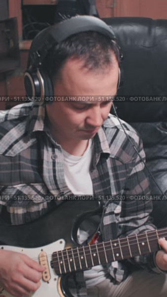 A musician plays an electric guitar while wearing headphones in a cozy, dimly lit room. The plaid shirt adds a casual vibe to the creative atmosphere. Стоковое видео, видеограф Воловодов Владимир / Фотобанк Лори